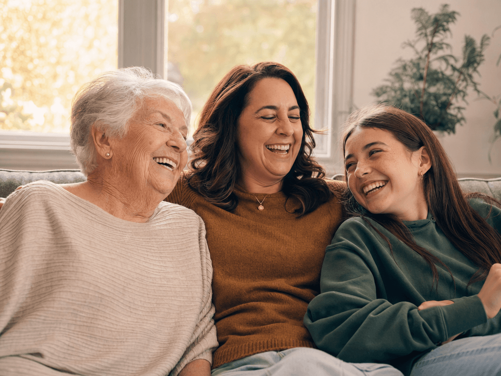 Grandmother, her daughter, and her granddaughter laughing together on a sunlit living room couch